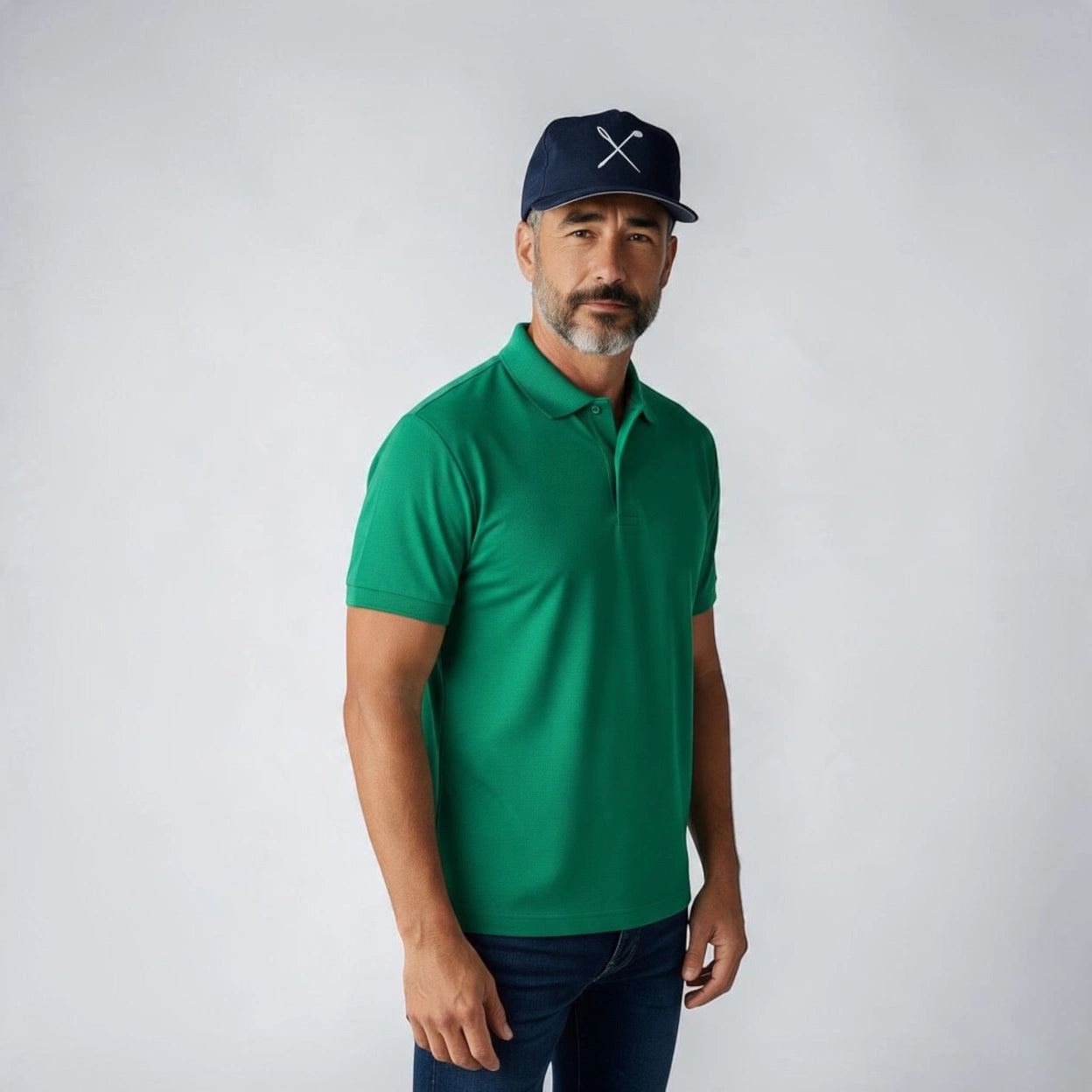 A man wearing a bright green short-sleeve polo shirt, dark blue jeans, and a navy blue baseball cap with a white crossed golf clubs logo, standing against a light gray studio background.
