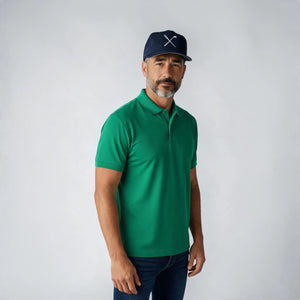 A man wearing a bright green short-sleeve polo shirt, dark blue jeans, and a navy blue baseball cap with a white crossed golf clubs logo, standing against a light gray studio background.